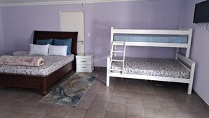 Quarto com cama de madeira, beliche branco, mesa de cabeceira pequena, televisão e uma porta.