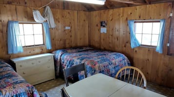 A cabin-style room with two beds, a dresser, a table, and two windows with curtains.