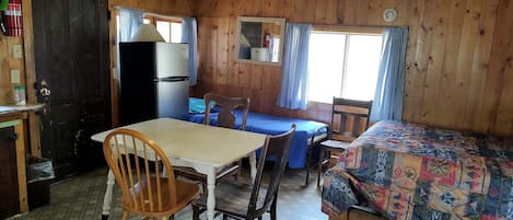 A wooden cabin with a dining table, chairs, a bed, a refrigerator, and a window with curtains.