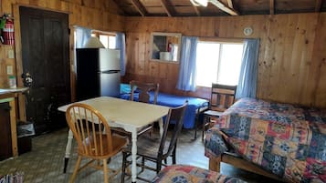 A wooden cabin with a dining table, chairs, a bed, a refrigerator, and a window with curtains.