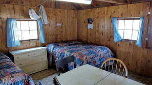 Habitación tipo cabaña con dos camas, un aparador, una mesa y dos ventanas con cortinas.