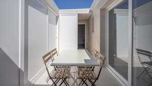 Un pati exterior modern amb taula blanca i cadires de fusta, grans portes de vidre i un cel blau clar.
