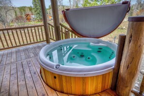 Ein Whirlpool auf einer Holzterrasse mit Holzgeländer, umgeben von Bäumen und einem Haus in der Ferne.