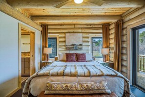 A log cabin bedroom with a bed, two bedside tables, a nightstand, a window, and a door leading to another room. - Hot Tub River Front Log Cabin (Pagosa Springs)