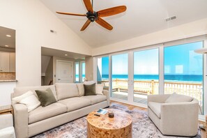 A spacious living area with a large window overlooking a beach, a ceiling fan, and a round coffee table. - Seascape - 4BR, Oceanfront, Dog Friendly, Fully Renovated! (Topsail Beach)