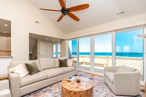 A spacious living room with a large window overlooking the beach, a ceiling fan, and a wooden coffee table. - Seascape - 4BR, Oceanfront, Dog Friendly, Fully Renovated! (Topsail Beach)