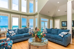 A spacious living room with a large window overlooking the ocean, a blue sofa set, a wicker coffee table, and a tropical plant centerpiece. - Sea Escape - Incredible Oceanfront 8 Bedroom Home in Gated Community (North Topsail Beach)