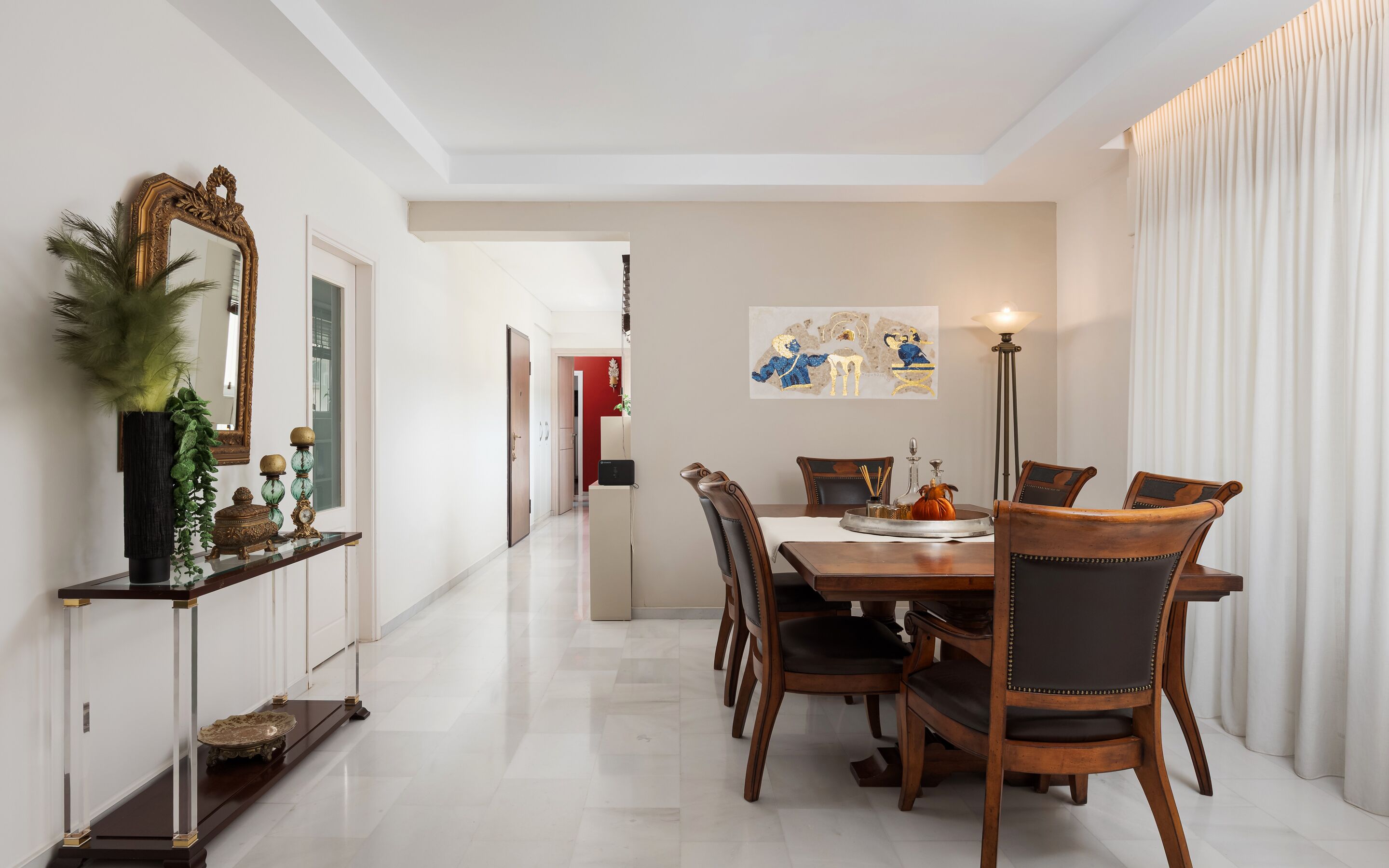 A dining area with a wooden table and chairs, a vase with green plants, a mirror, and a painting on the wall.
