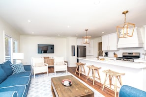 A modern living room with a sofa, coffee table, and a kitchen area with white cabinets and a stove. - The Canady Cottage- 3 BR Home Located on a Quiet Street (Topsail Beach)