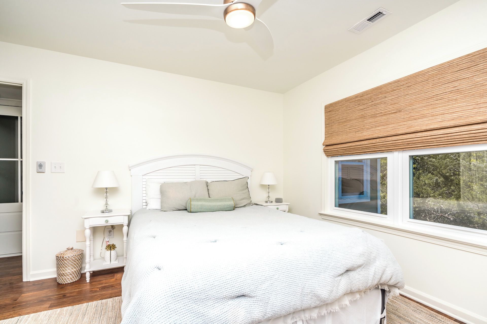 A bedroom with a bed, bedside tables, a ceiling fan, a window with a view, and a door.