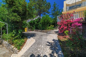 Ein gepflasterter Weg führt zu einem Gebäude mit einem Balkon, umgeben von üppiger Vegetation und blühenden Pflanzen.
