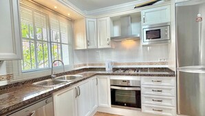 A modern kitchen with white cabinetry, a stainless steel refrigerator, a built-in oven, a microwave, and a window with blinds. - TERRASOL LOS NARDOS 229 (Caleta de velez)