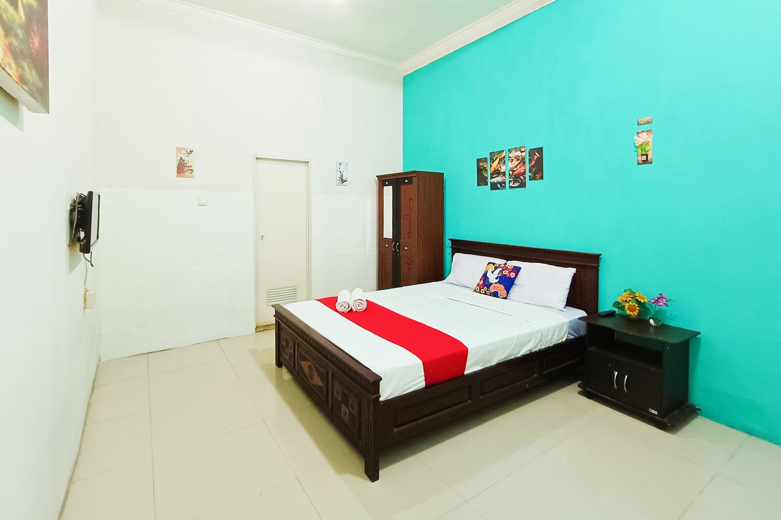 A bedroom with a wooden bed, a red bedspread, a nightstand with a vase of flowers, and a teal-colored wall.