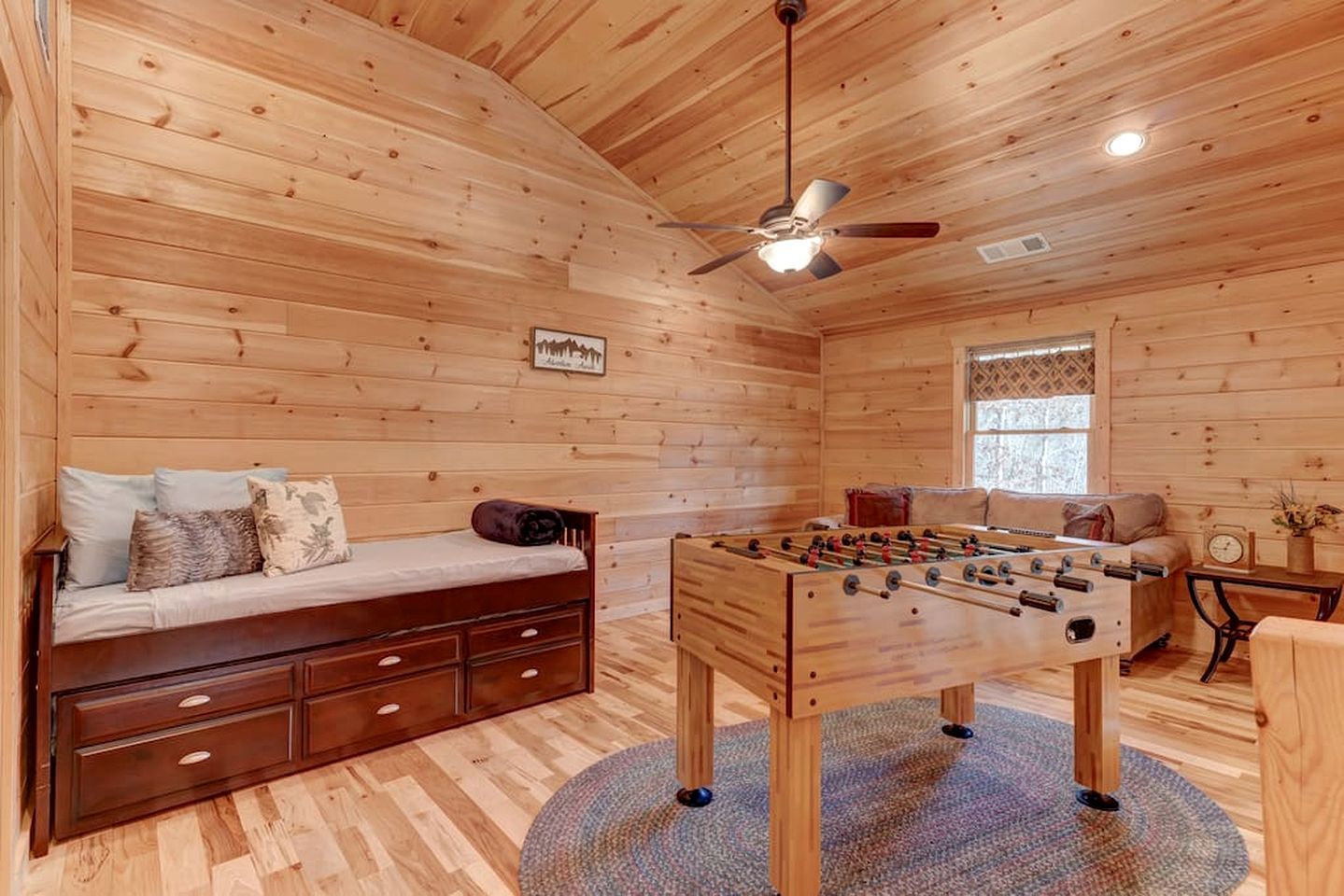 A cozy room with a wooden ceiling, a ceiling fan, a sofa with cushions, a foosball table, and a window with patterned curtains.