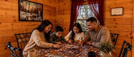 Ein Holzhütten-Innenraum, in dem eine Familie ein Puzzlespiel spielt, ein gerahmtes Gemälde hängt und ein Schild mit einem Bibelvers angebracht ist.