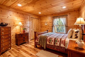 Ein Schlafzimmer in einer Holzhütte mit Bett, Kommode und einem fenster mit Vorhängen.