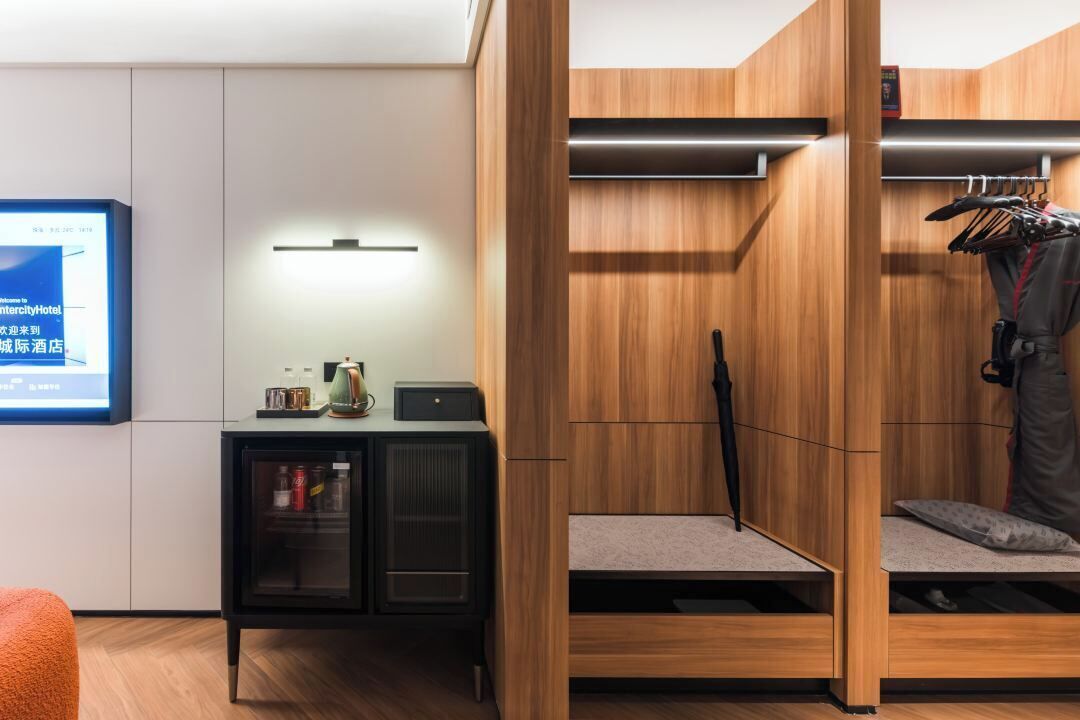 A modern hotel room with a wooden wardrobe, a small desk with a lamp, and a bed with a pillow.