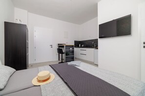 A modern bedroom with a bed, a hat, a TV, a kitchen area, and a white door. - Modern studio with sea view & balcony in Crikvenica (Crikvenica)