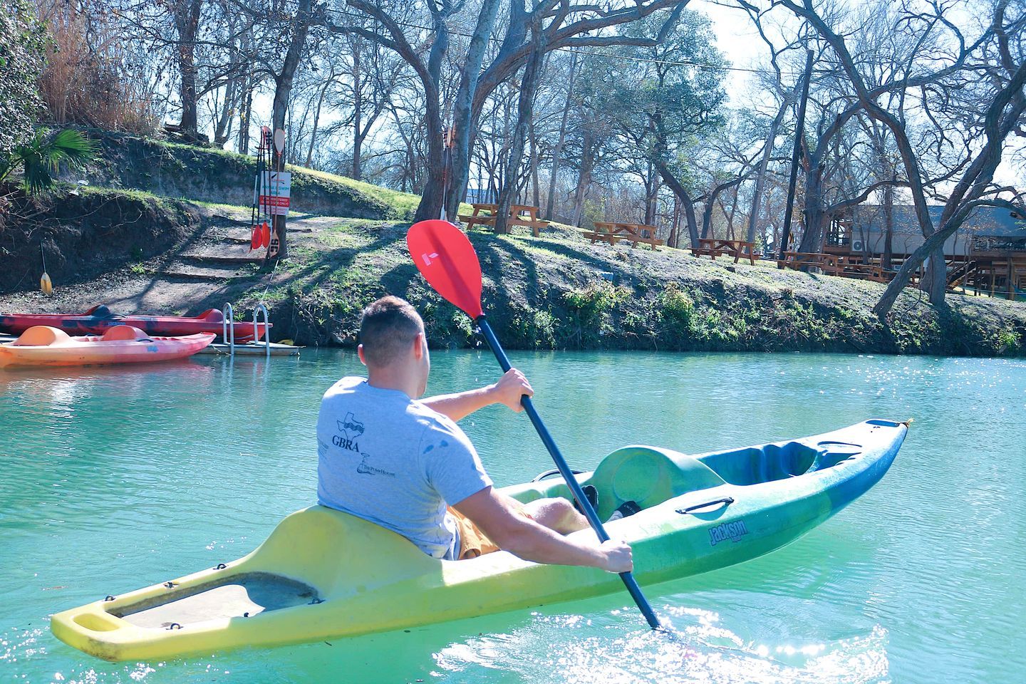 Una persona en un kayak sobre un río tranquilo, rodeada de árboles y mesas de picnic.