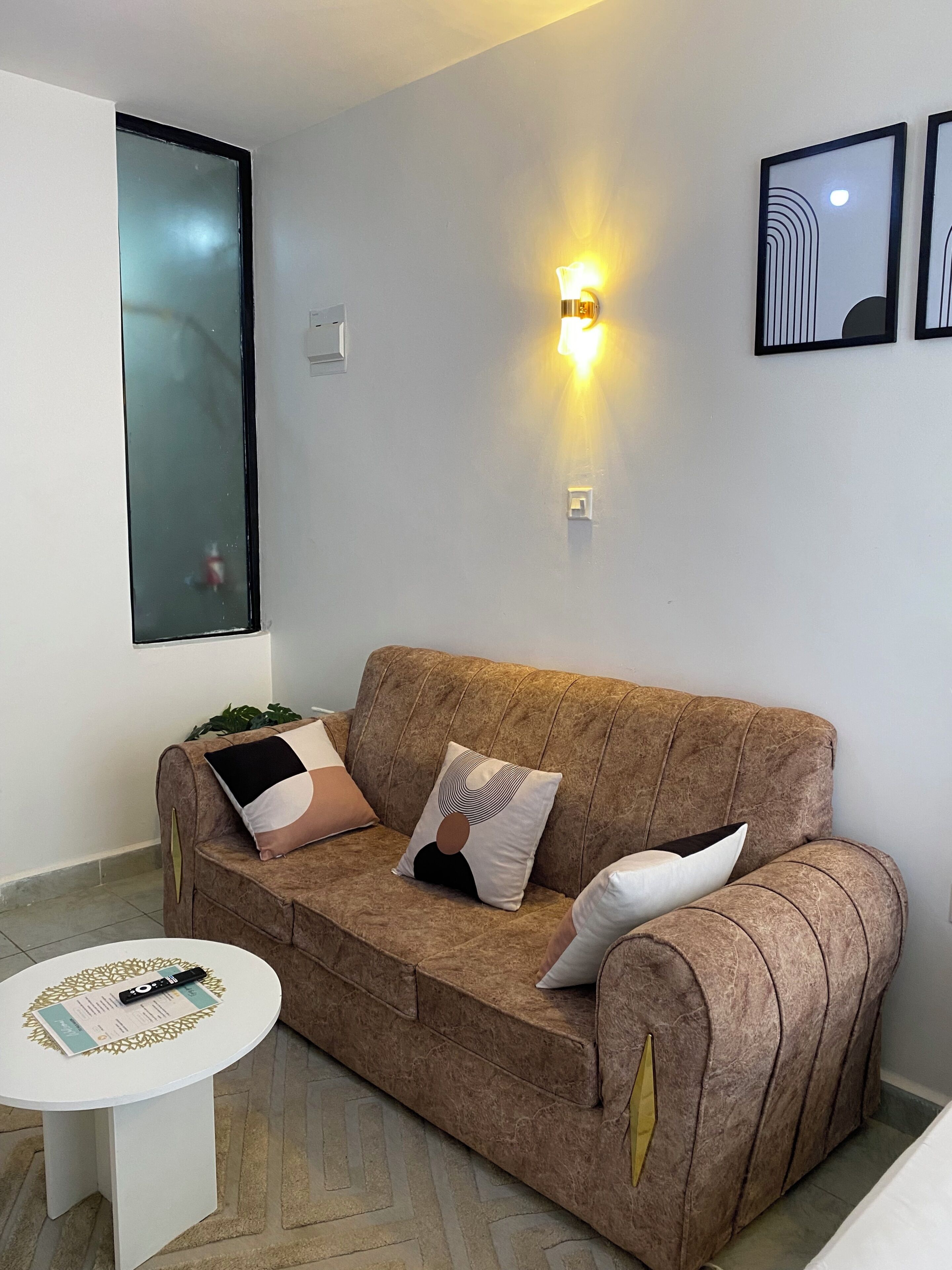 A brown sofa with patterned cushions, a small white table with a document, and a wall-mounted light fixture.