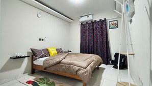Een slaapkamer met een bed, nachtkastje, airconditioning aan de muur, raam met gordijnen en een klein plankje met toiletartikelen.