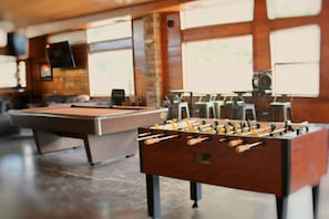 A billiards table and a foosball table in a room with large windows and a brick wall.