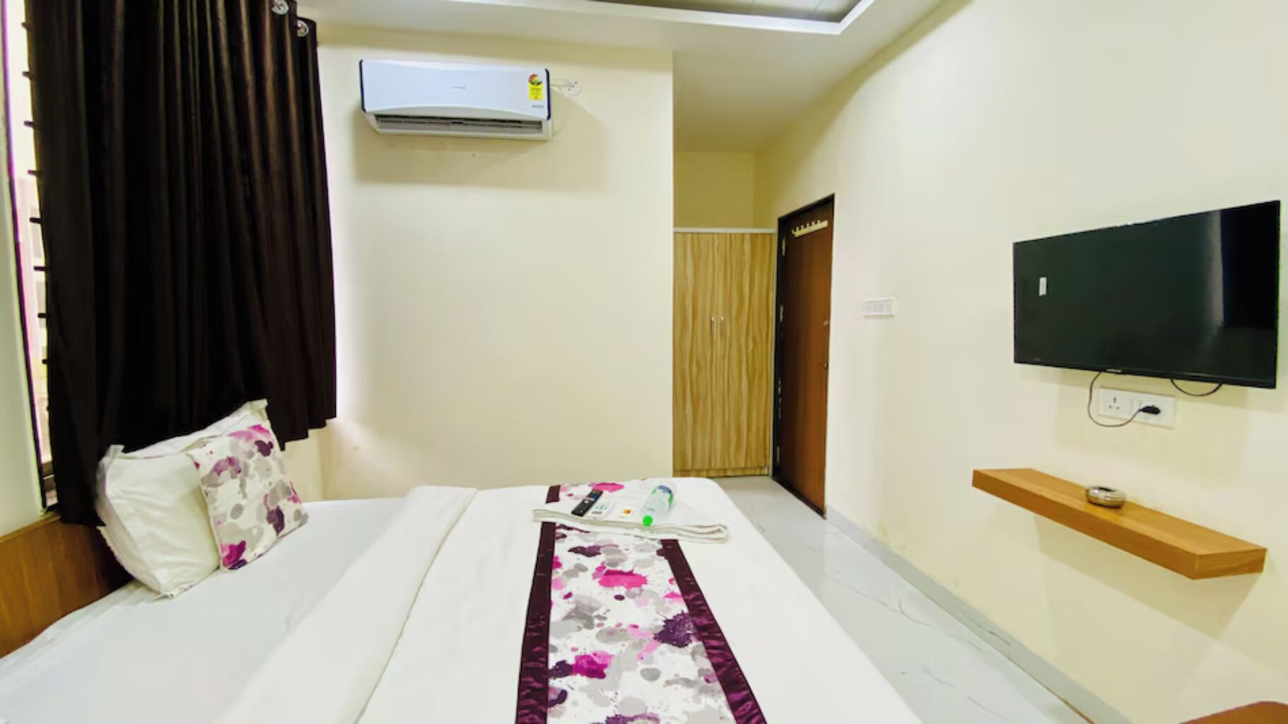 A hotel room with a bed, a TV, an air conditioner, a wooden door, and a small shelf.