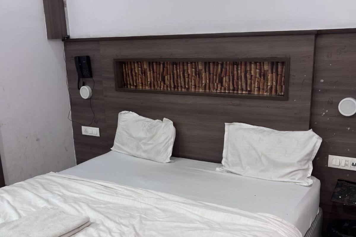 A neatly made bed with white linens, a wooden headboard with a cork panel, a bedside telephone, and a power outlet on the wall.