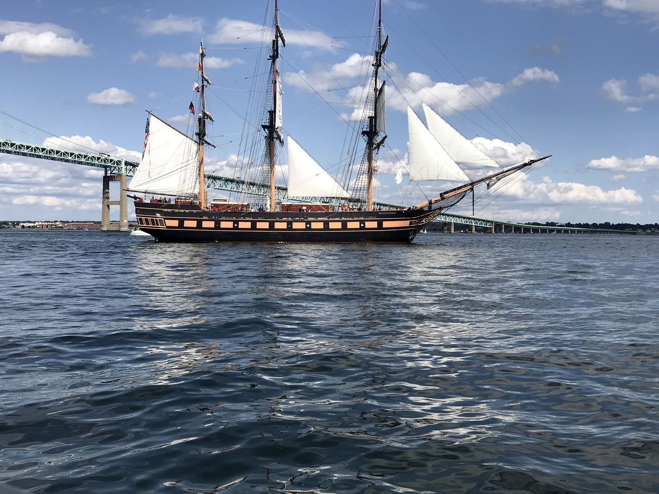 A large sailing ship with multiple masts and sails, docked near a bridge.