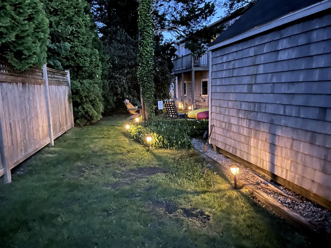 A backyard with a wooden fence, a house, and a pathway lit by ground lights.
