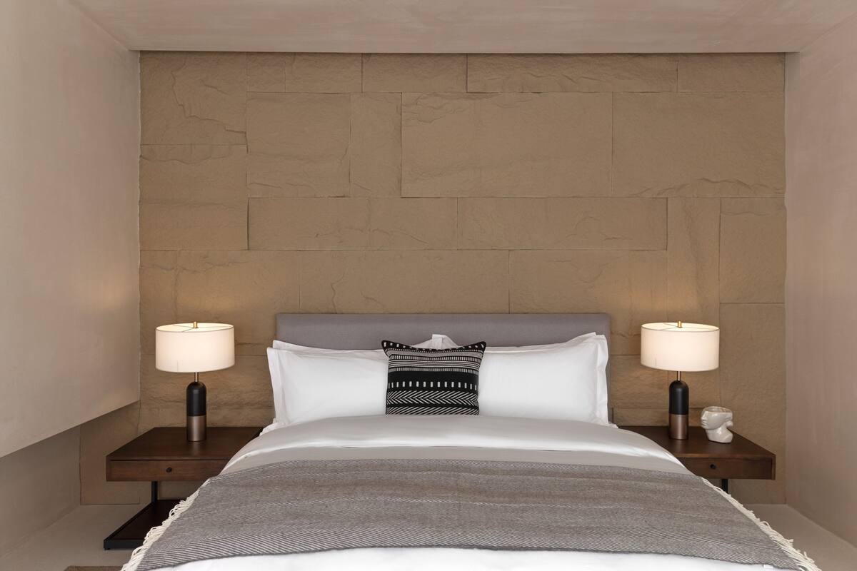 Ein ordentlich bezogenes Bett mit weißen Leinen, zwei Nachttische mit Lampen, ein gemustertes Kopfkissen und eine strukturierte Wand dahinter.