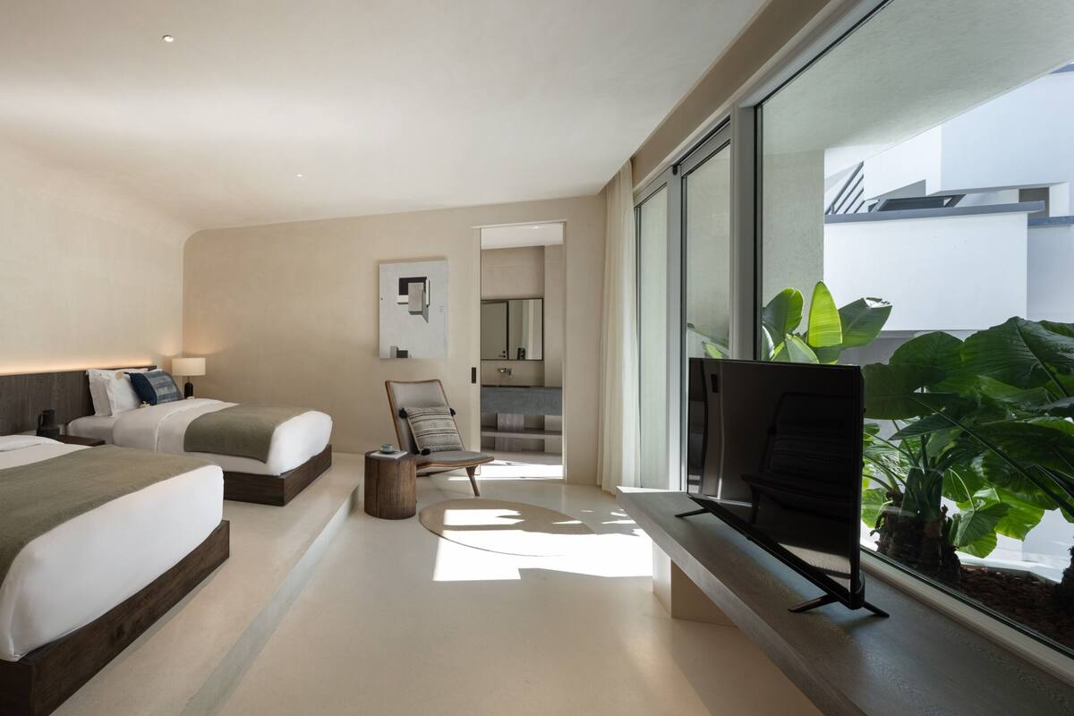 Ein modernes Hotelzimmer mit zwei Betten, einem Fernseher, einem großen Fenster mit Aussicht und einer kleinen Sitzecke.