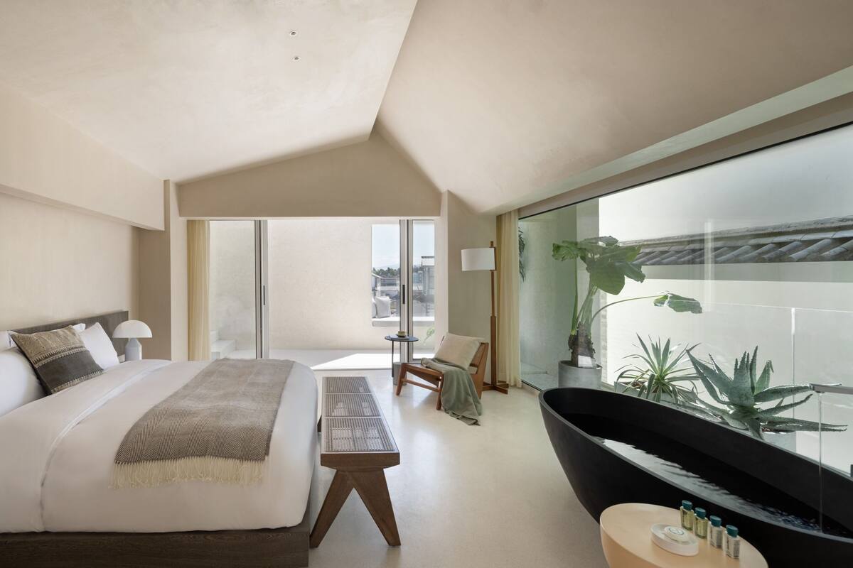 Ein modernes Hotelzimmer mit einem großen Bett, einer Sitzecke mit Tisch und Stühlen, einer Badewanne und Blick auf die Stadt.