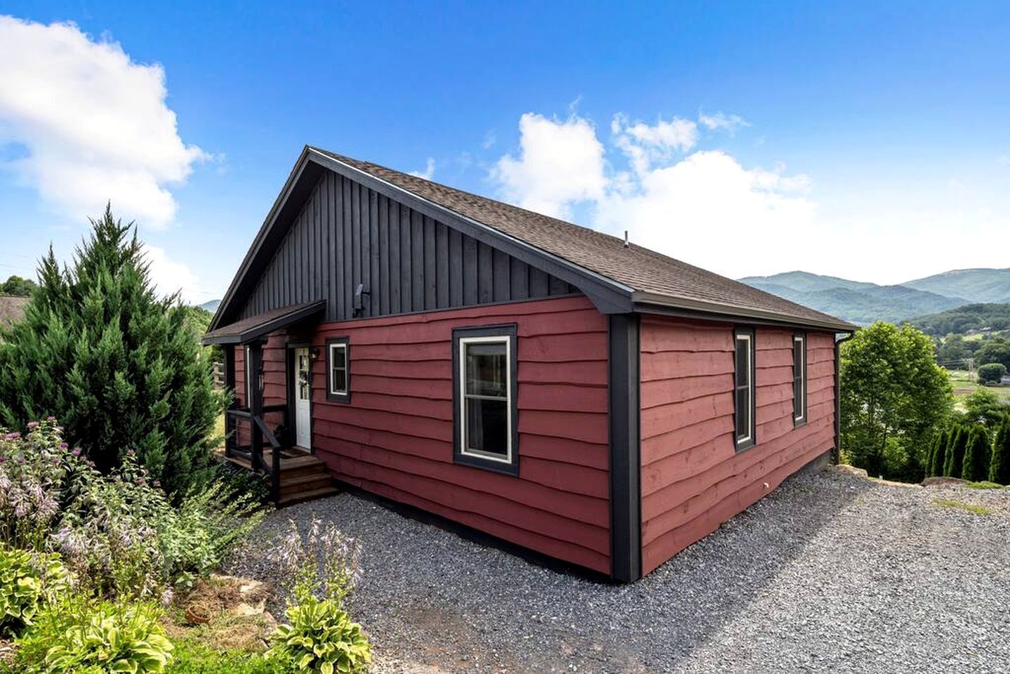 Une petite maison de plain-pied, avec un toit sombre et un revêtement rouge, entourée d’une allée en gravier et d’une végétation luxuriante.