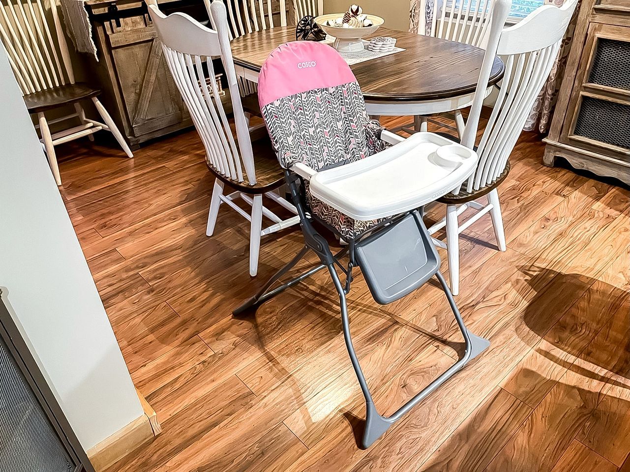 A dining area with a round table, white chairs, and a high chair with a patterned seat.
