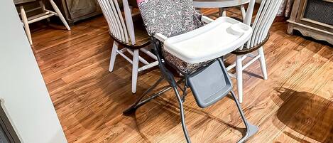 A dining area with a round table, white chairs, and a high chair with a patterned seat.