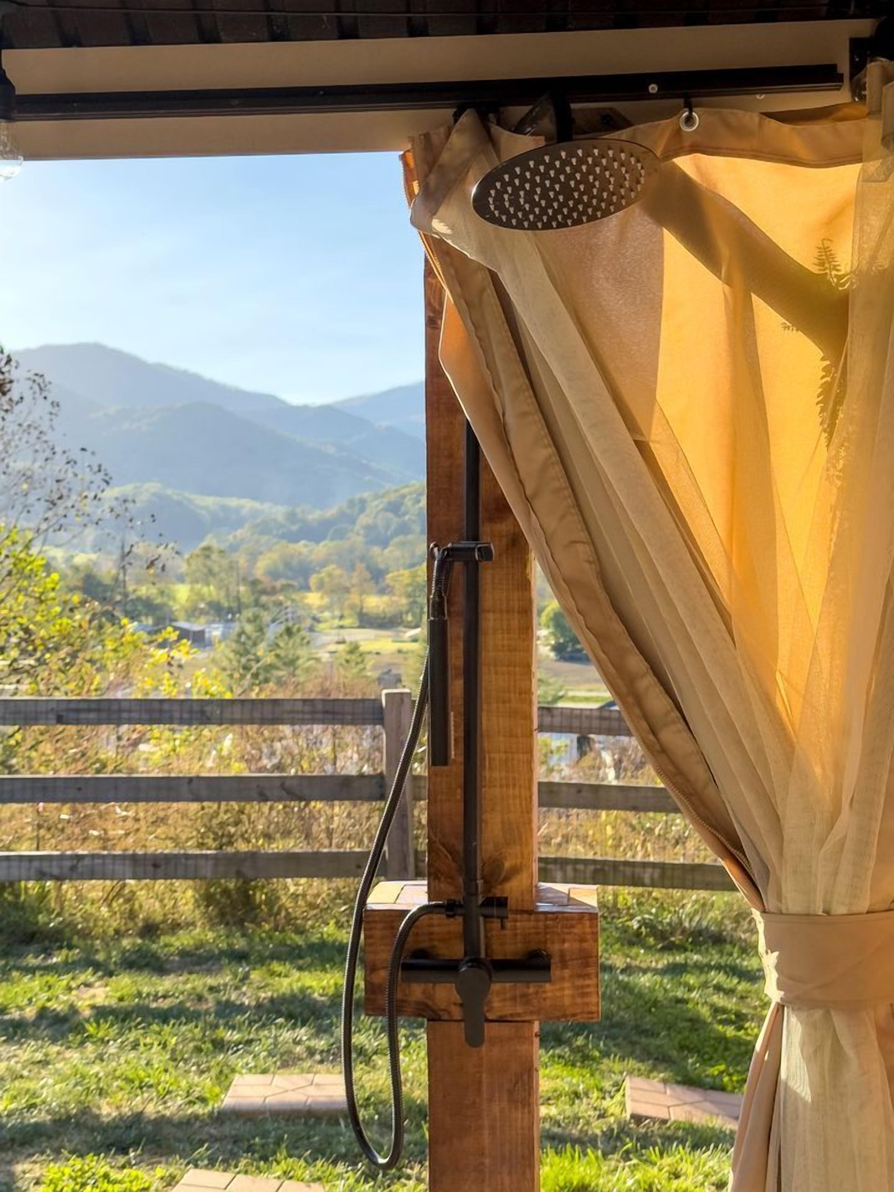 Une douche extérieure en bois, équipée d’un tuyau d’arrosage, d’un rideau et offrant une vue sur un paysage montagneux.
