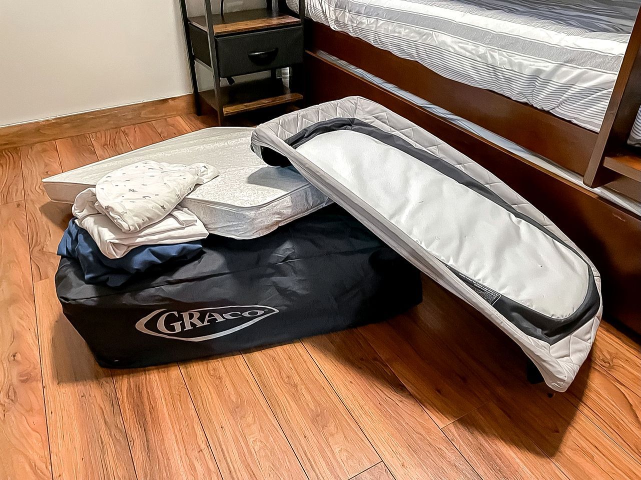 A room with a bed, a wooden floor, a black GRACO suitcase, and a white mattress.