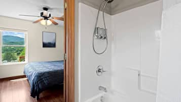 A bedroom with a bed, a ceiling fan, a window with a view, and a bathroom with a shower.
