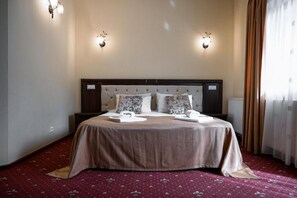 A hotel room with a large bed, two bedside tables, a headboard with light fixtures, and a window with curtains.