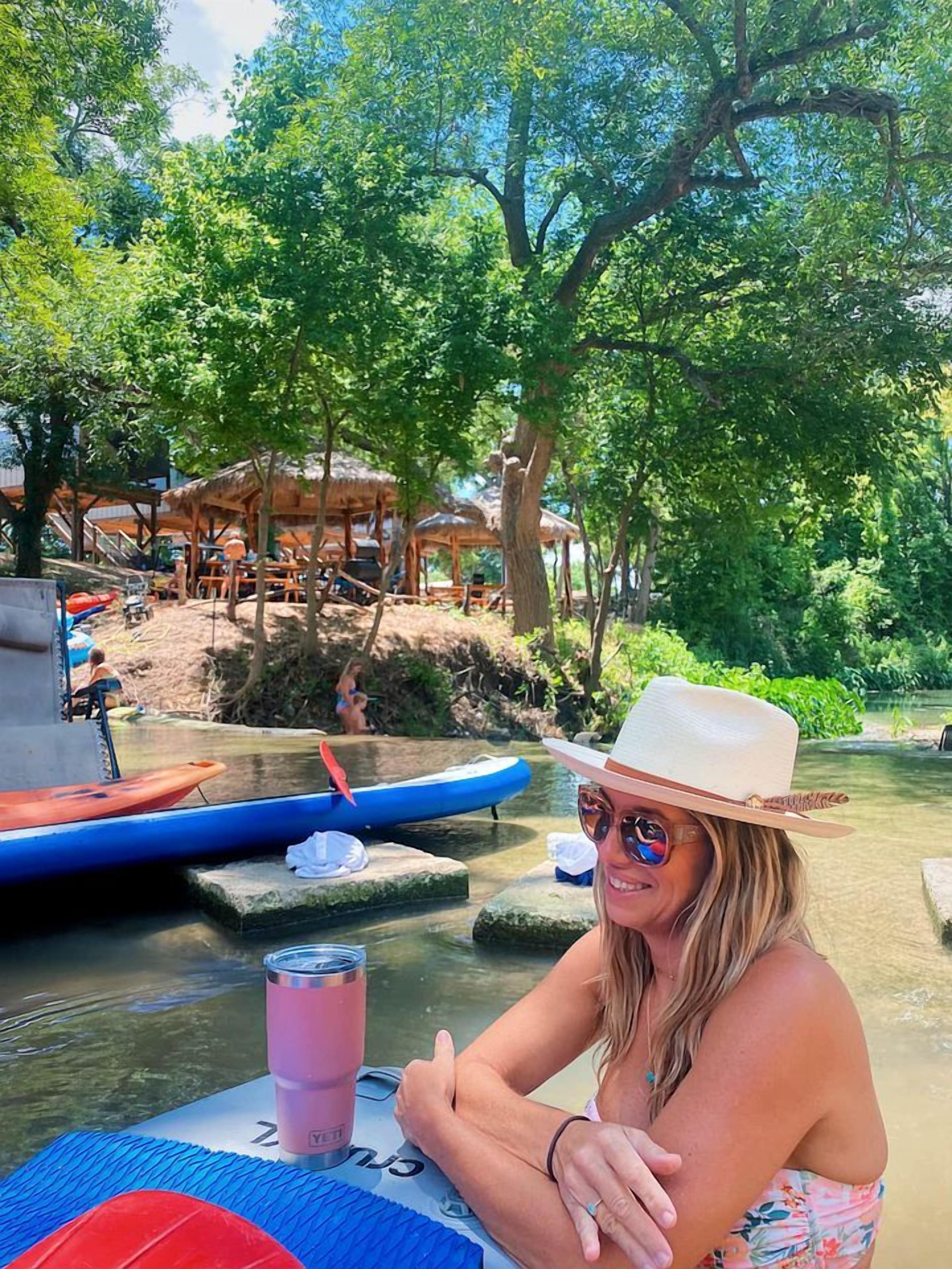 A person seated at a table with a drink, surrounded by a natural setting with trees and a water body.