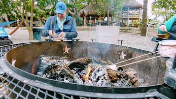 A person grilling outdoors with a fire pit, surrounded by trees and a thatched-roof structure.
