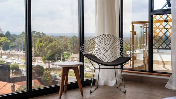 A modern room with a large window, a metal-framed chair, a small wooden stool, and a view of a cityscape. - ORAK - Minimalist Aparthotel with Gym & Pool in Lavington (Nairobi)
