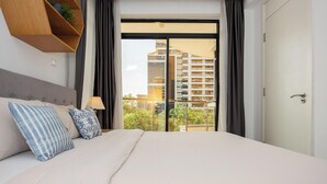 A hotel room with a bed, a nightstand, a lamp, a window with curtains, and a view of a building. - ORAK - Minimalist Aparthotel with Gym & Pool in Lavington (Nairobi)