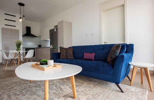 A modern living room with a blue sofa, a white coffee table, and a kitchen in the background. - ORAK - Minimalist Aparthotel with Gym & Pool in Lavington (Nairobi)