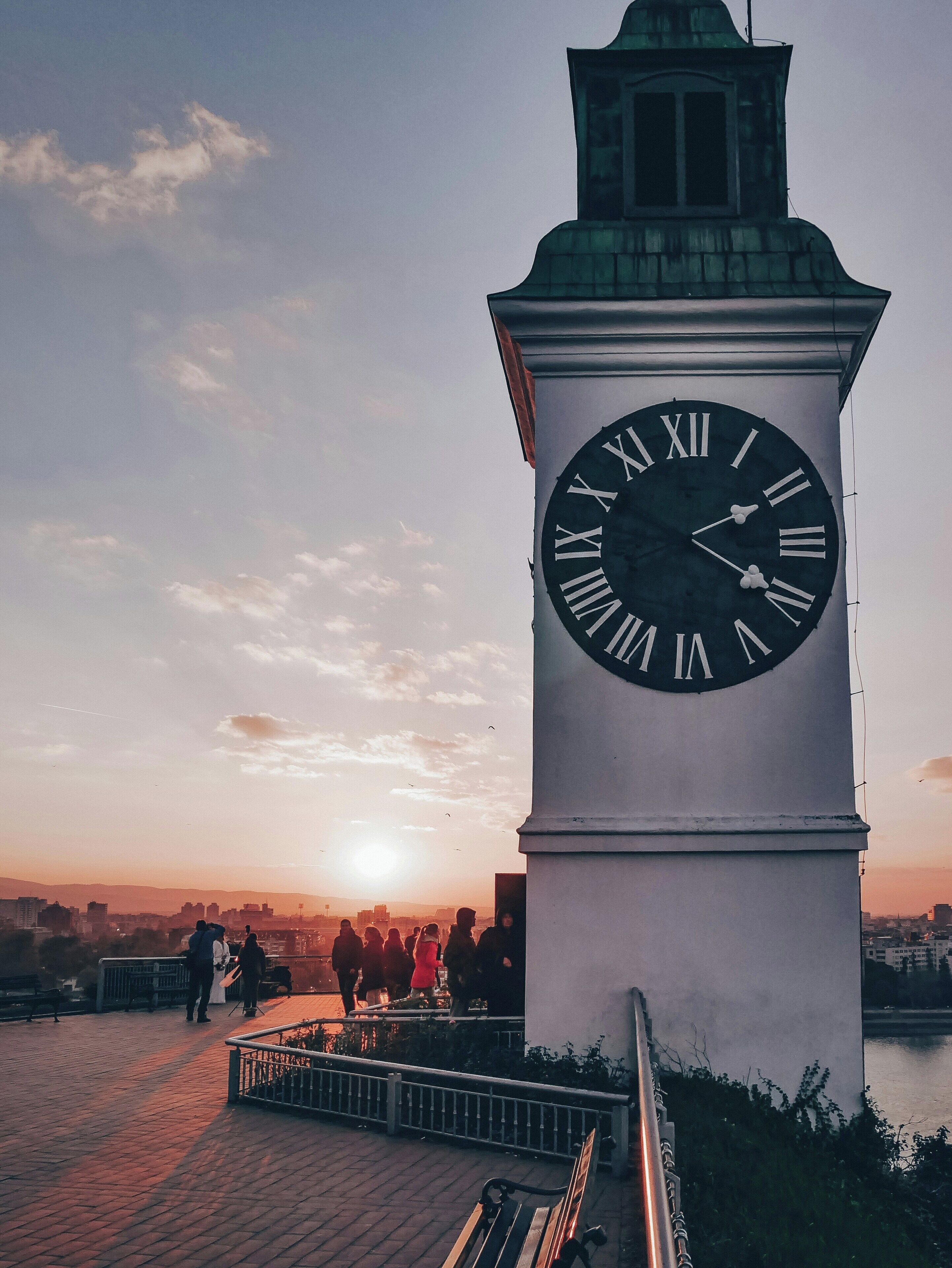 Una torre del reloj con números romanos, un camino empedrado y una vista del paisaje urbano al fondo.