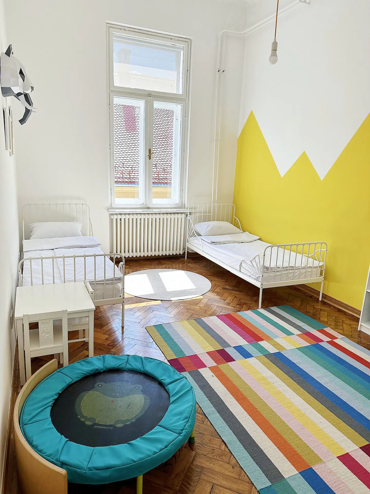 Een kinderkamer met twee bedden, een klein tafeltje, een kleurrijk tapijt, een trampoline en een raam met uitzicht op een gebouw.