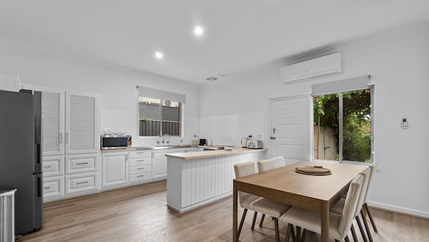 A modern kitchen with white cabinetry, a dining area with a wooden table and chairs, and a large window providing a view of greenery outside.