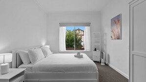 A white bedroom with a bed, bedside tables, a window with curtains, a radiator, and a wall-mounted artwork.