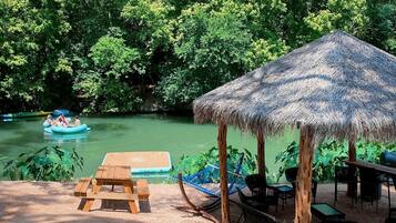 A riverside area with a thatched-roof pavilion, picnic tables, and lounge chairs.